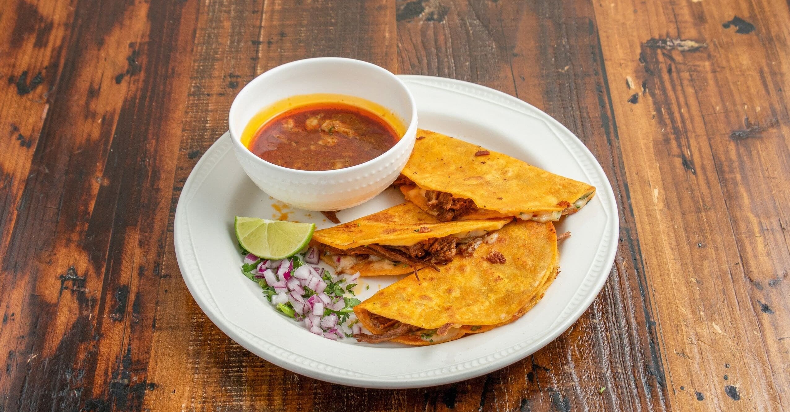 Three crispy quesabirria tacos with consommé, lime, onion, and cilantro on a white plate