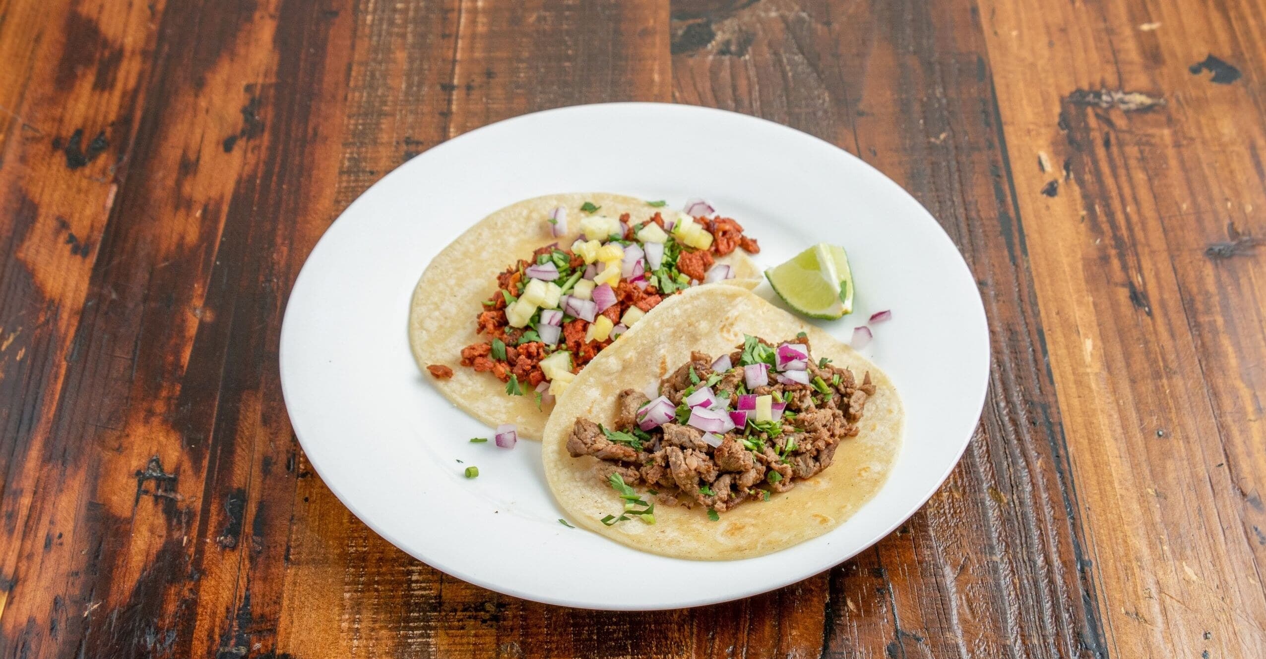Two street tacos with al pastor and carne asada, topped with onion, cilantro, and a lime wedge